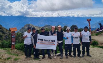 A group of tourists holding a Kenko Adventures flag in the Sacred Valley of the Incas, with terraced hills and towering mountains in the background. A group of tourists holding a Kenko Adventures flag in the Sacred Valley of the Incas, with terraced hills and towering mountains in the background.